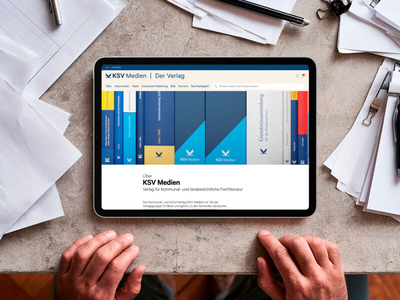 Auf einem iPad wird die Über-Seite des KSV Medien Webshops angezeigt. Das iPad liegt auf einer Tischplatte, inmitten von Papieren und Utensilien. Unten am Tischrand liegen zwei Hände auf.